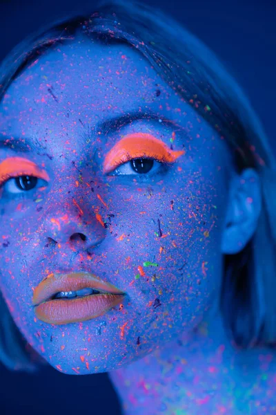 Nahaufnahme Porträt einer Frau in bunten Farbspritzern und Neon-Make-up isoliert auf dunkelblau — Stockfoto