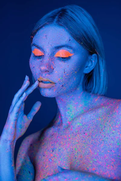Mulher sensual com maquiagem de néon e tinta brilhante no corpo tocando lábios isolados em azul escuro — Fotografia de Stock
