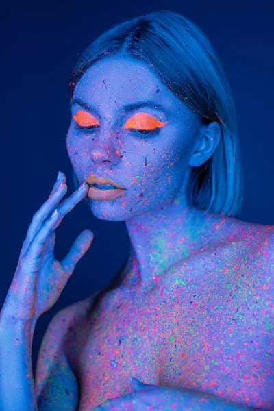 Mujer sensual con maquillaje de neón y pintura brillante en el cuerpo tocando los labios aislados en azul oscuro — Stock Photo