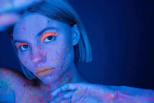 Портрет женщины с флуоресцентным макияжем и красочной неоновой краской тела на размытом переднем плане, изолированном на темно-синем — стоковое фото