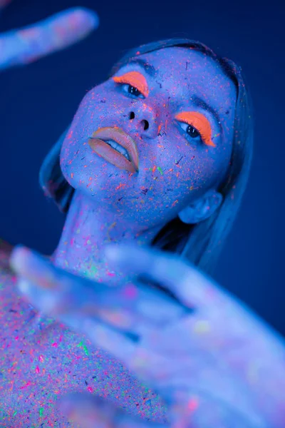 Mujer sensual con maquillaje de neón brillante y pintura fluorescente posando en primer plano borroso aislado en azul oscuro - foto de stock