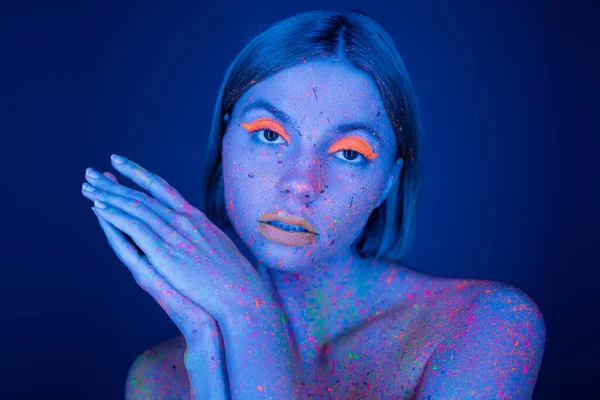 Hübsche Frau in Neon-Make-up und bunter Farbe, die isoliert auf dunkelblau in die Kamera blickt — Stockfoto