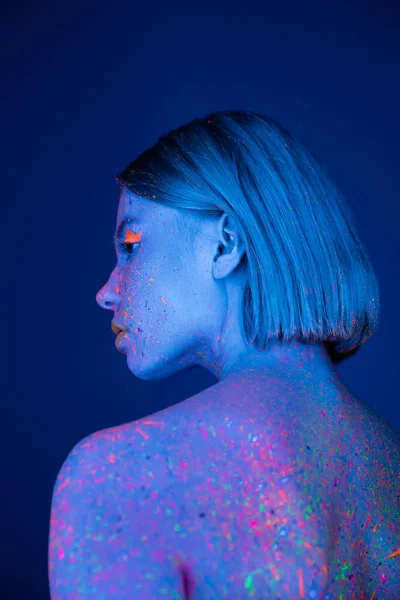 Rückansicht einer jungen Frau in leuchtendem Make-up und heller Neon-Körperfarbe isoliert auf dunkelblau — Stockfoto