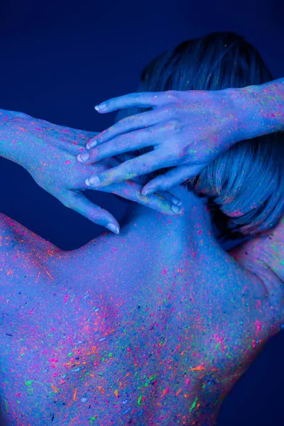 Rückansicht einer Frau in leuchtender Neon-Körperfarbe, die mit den Händen hinter dem Kopf isoliert auf dunkelblauem Hintergrund posiert — Stockfoto