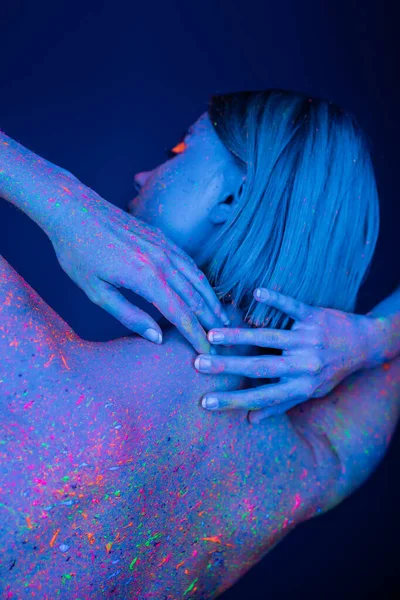 Rückansicht einer jungen Frau in leuchtender Neonfarbe, die die Hände hinter dem Hals hält, isoliert auf dunkelblau — Stockfoto