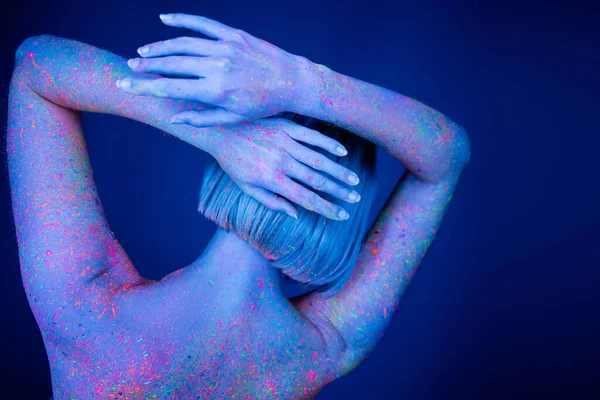 Vue arrière de la femme en peinture fluorescente au néon debout avec les mains derrière la tête isolée sur bleu foncé — Photo de stock