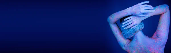 Vue arrière de la femme colorée avec de la peinture au néon posant avec les mains derrière la tête isolée sur bleu foncé, bannière — Photo de stock