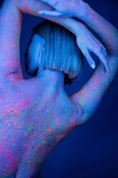 Vista posterior de mujer desnuda coloreada con pintura de neón posando con las manos detrás de la cabeza aisladas en azul oscuro - foto de stock