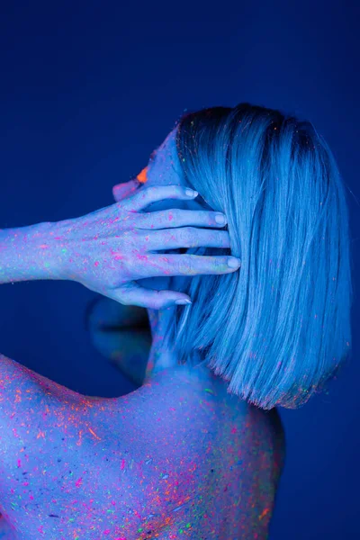 Young woman colored with neon paint touching hair isolated on dark blue — Stock Photo