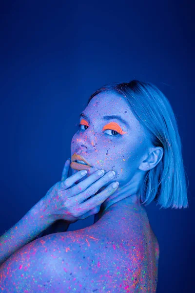 Giovane donna con vibranti neon visage e corpo colorato toccare il viso e guardando la fotocamera isolata su blu scuro — Foto stock