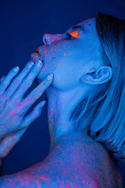 Vista laterale della donna nel trucco al neon e vernice per il corpo che tiene le mani vicino al viso isolato sul blu scuro — Foto stock