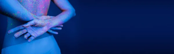 Partial view of woman with body in neon paint standing with hands behind back isolated on dark blue, banner — Stock Photo