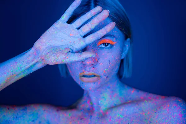 Mulher em maquiagem de néon e rosto de obscurecimento de tinta corporal brilhante com a mão enquanto olha para a câmera isolada no azul escuro — Fotografia de Stock