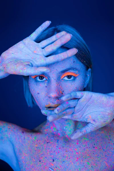 Jeune femme en maquillage fluorescent et peinture au néon brillant tenant les mains près du visage isolé sur bleu foncé — Photo de stock