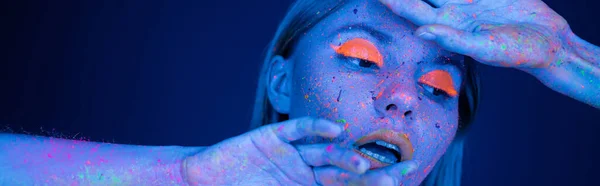 Donna sensuale con vibrante trucco al neon posa con le mani vicino al viso isolato su blu scuro, banner — Foto stock