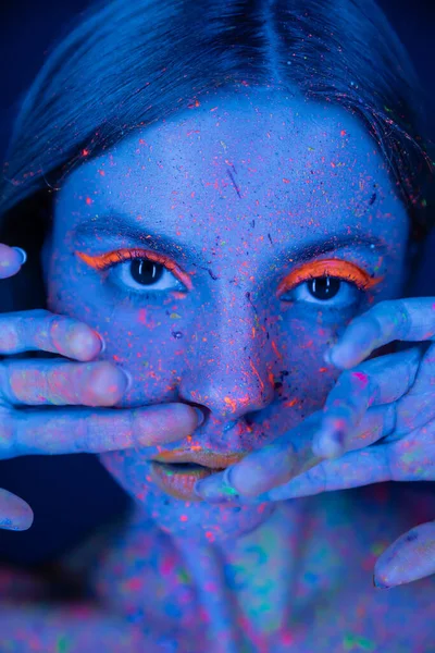 Portrait of woman holding hands near face with neon makeup and fluorescent paint isolated on dark blue — Stock Photo