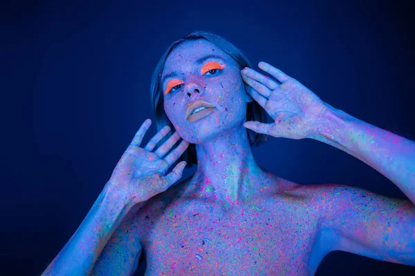 Nackte Frau in Neon-Make-up und Körper in glühender Farbe posiert mit Händen in Gesichtsnähe isoliert auf dunkelblau — Stockfoto