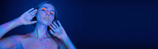 Jeune femme avec maquillage au néon et éclaboussures de peinture brillantes sur le corps en regardant la caméra isolée sur bleu foncé, bannière — Photo de stock