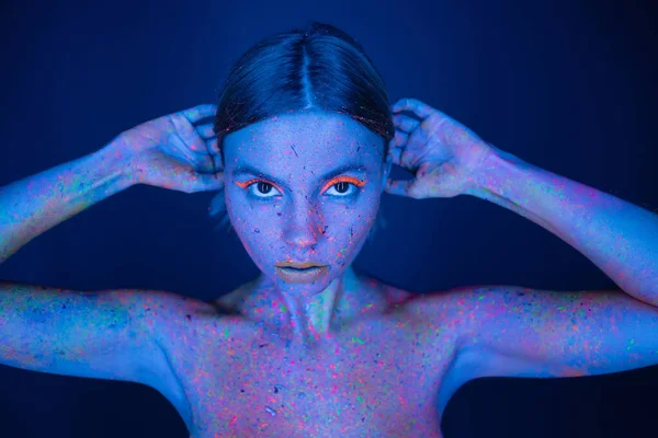 Mujer desnuda en brillante pintura de neón y maquillaje brillante posando con las manos detrás de la cabeza aisladas en azul oscuro - foto de stock