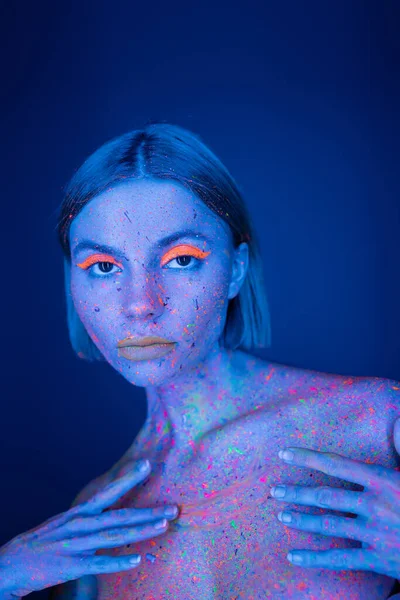 Retrato de mulher com maquiagem de néon brilhante tocando corpo colorido isolado em azul escuro — Fotografia de Stock