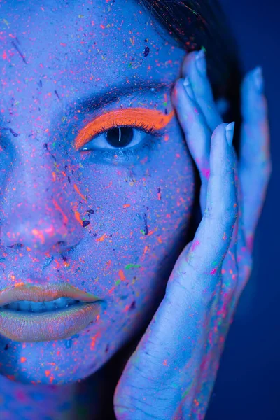 Close up portrait of cropped woman touching face with neon makeup and vibrant paint splashes isolated on dark blue — Stock Photo