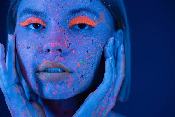 Porträt einer Frau in leuchtendem Neon-Make-up und Körperfarbe, die Gesicht berührt und isoliert auf dunkelblau in die Kamera blickt — Stockfoto