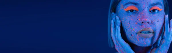 Ritratto di giovane donna che tocca il viso con trucco al neon e spruzzi di vernice fluorescente isolati su blu scuro, banner — Foto stock