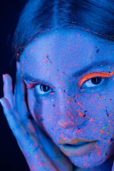 Nahaufnahme Porträt einer Frau mit Neon-Make-up und bunter Farbe im Gesicht, die isoliert auf dunkelblau in die Kamera blickt — Stockfoto