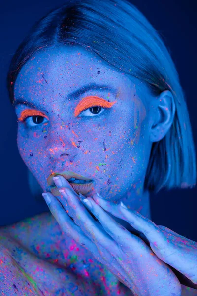 Портрет жінки в яскравому неоновому макіяжі і барвиста фарба для тіла, що торкається губ ізольовані на темно-синьому — стокове фото