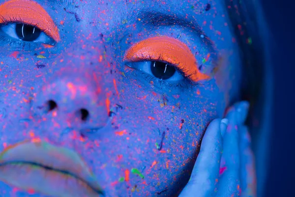 Close up view of cropped woman touching face with neon makeup and bright paint splashes in blue light — Stock Photo