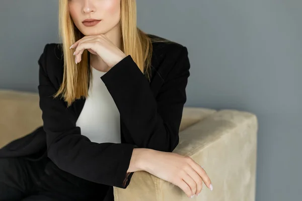 Vue recadrée de la femme blonde en veste noire assise sur le canapé près du mur gris — Photo de stock