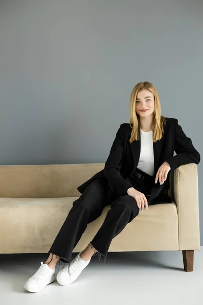 Усміхнена блондинка в джинсах і чорна куртка сидить на дивані вдома — стокове фото