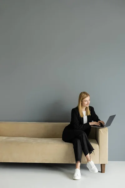 Smiling blonde coach using laptop while sitting on beige sofa at home — Stock Photo