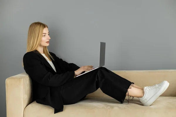 Coach blonde utilisant un ordinateur portable assis sur un canapé beige à la maison — Photo de stock
