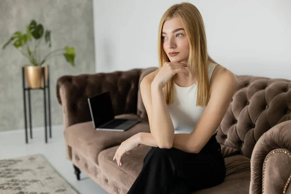 Jeune femme blonde en vêtements décontractés regardant loin près d'ordinateur portable flou sur le canapé — Photo de stock