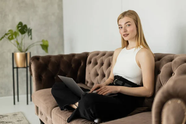 Lächelnde blonde Frau im Top mit Laptop und Blick auf die Kamera auf der Couch — Stockfoto