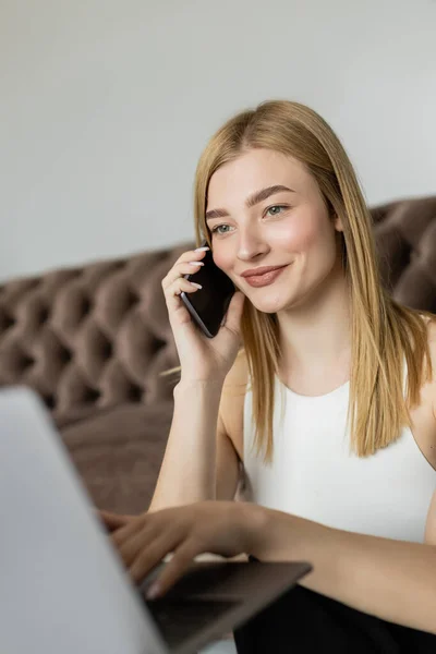 Portrait of smiling blonde coach talking on cellphone and using blurred laptop in living room — Stock Photo