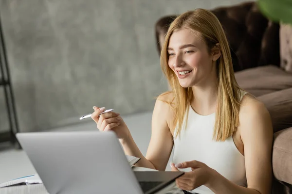 Femme blonde souriant tout en tenant un stylo près d'un ordinateur portable et des ordinateurs portables pendant l'éducation en ligne — Photo de stock