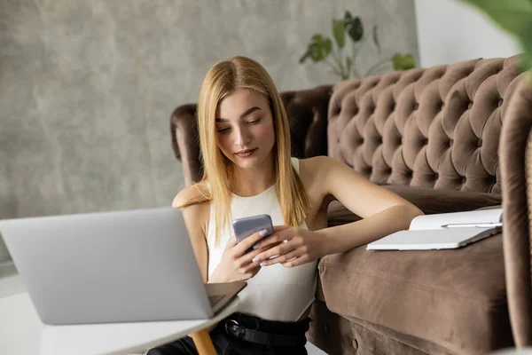 Hübsche Blondine nutzt Smartphone in der Nähe von Laptop und Notebook auf der Couch — Stockfoto