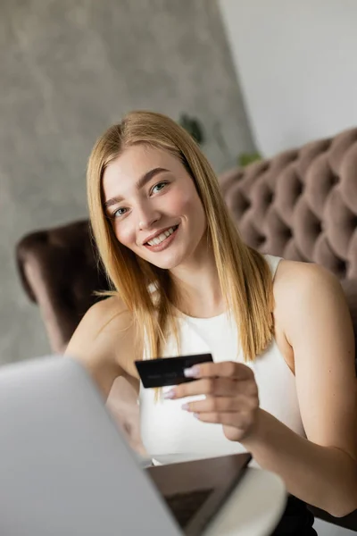 Mulher loira feliz segurando cartão de crédito perto de laptop borrado e olhando para a câmera em casa — Fotografia de Stock