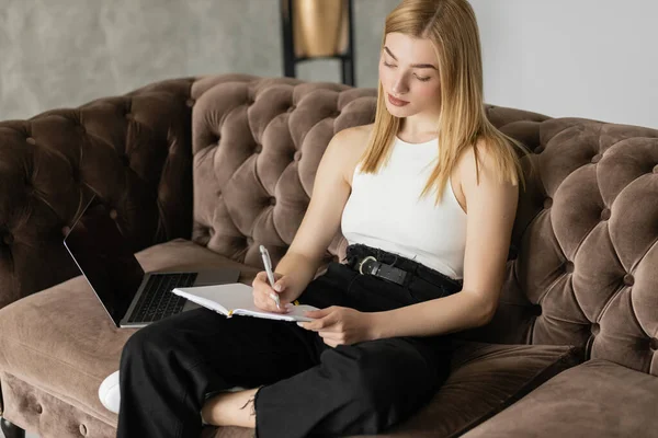 Young blonde woman writing on notebook during e-learning on laptop on couch — Stock Photo