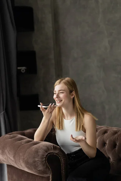 Smiling blonde woman recording voice message while sitting on armchair — Stock Photo