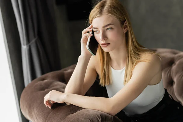 Сосредоточенная молодая женщина разговаривает по смартфону, сидя дома на кресле — стоковое фото