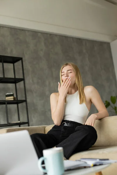 Freelance bionda sbadigliando vicino a gadget sfocati e caffè in soggiorno — Foto stock