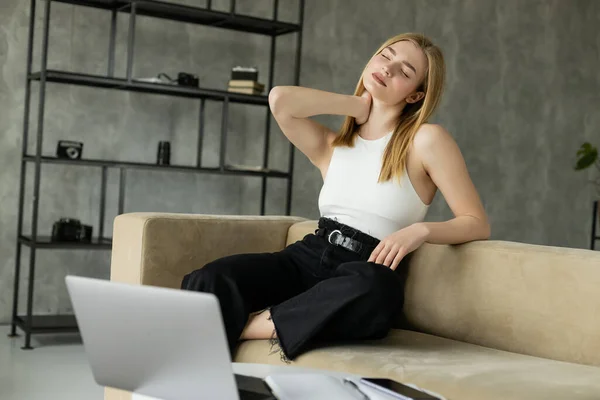 Exhausted blonde freelancer touching neck near devices on couch — Stock Photo