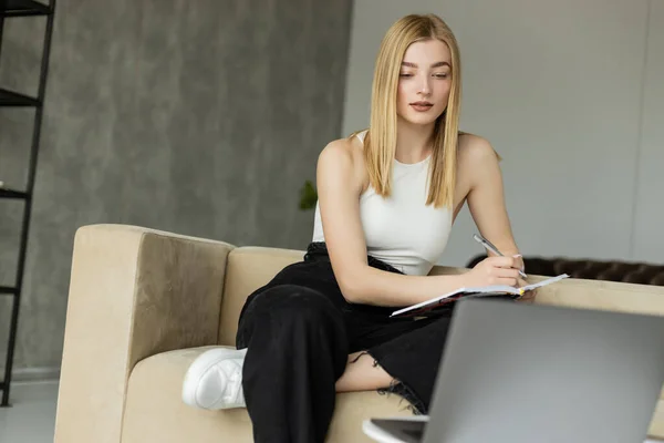 Young woman in top writing on notebook near blurred laptop during online education — Stock Photo