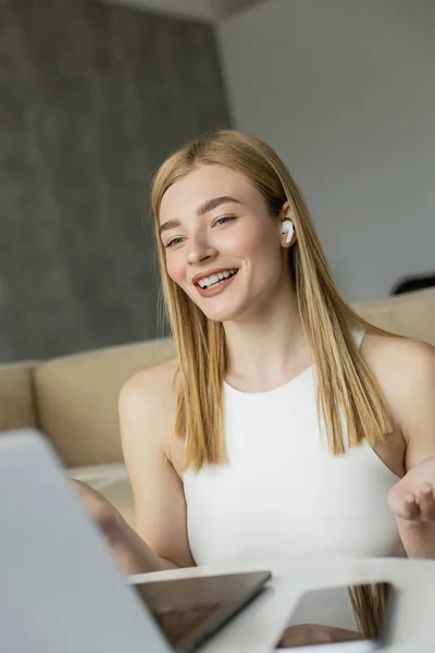 Positive blonde coach in earphone having video chat on blurred laptop in living room — Stock Photo