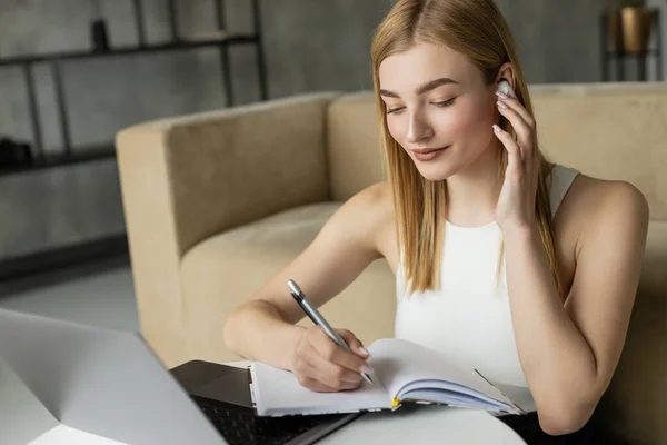 Femme blonde écoutant de la musique dans un écouteur et écrivant sur un portable près d'un ordinateur portable pendant l'éducation en ligne à la maison — Photo de stock