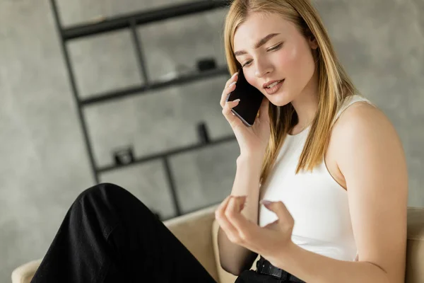 Jeune femme en vêtements décontractés regardant la main tout en parlant sur un téléphone portable dans le salon — Photo de stock