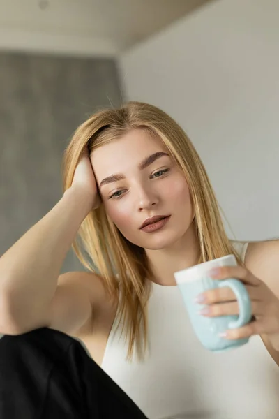 Thoughtful blonde woman holding blurred cup of coffee at home — Stock Photo